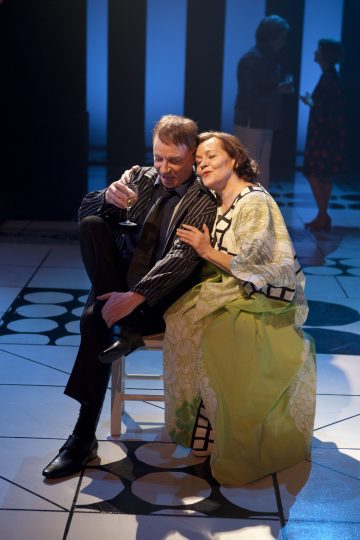 Pukumies, jolla on drinkki kädessään, istuu lähekkäin vihreään kuviomekkoon pukeutuneen naisen kanssa lavalla, ja molemmat hymyilevät hellästi. Taustalla sinisessä valaistuksessa keskustelevat kaksi epäselvää hahmoa.