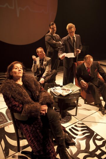 Viisi virallisesti pukeutunutta henkilöä on kokoontunut hämärään huoneeseen, jossa on dramaattinen valaistus. Etualalla istuu turkisturkkinen nainen, ja hänen takanaan seisoo tai istuu neljä miestä, jotka lukevat lehtiä ja keskustelevat.