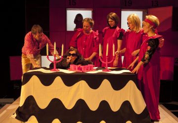 Kuusi kirkkaisiin vaaleanpunaisiin ja punaisiin asuihin pukeutunutta ihmistä seisoo pöydän ympärillä, jossa on mustavalkoinen aaltoileva pöytäliina. Yksi aurinkolaseja käyttävä henkilö nojaa pöydälle vaaleanpunaisten kynttilöiden viereen, kun taas toinen henkilö katsoo ikkunallisen taustan läpi.