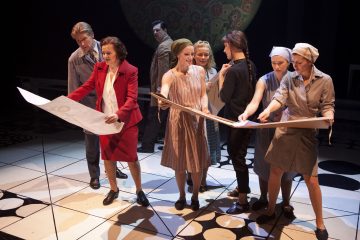 Ryhmä esiintyjiä tutkii lavalla suuria paperiarkkeja. Useimmat ovat 1950-luvun tyyliin pukeutuneita naisia, joilla on päähuivi, yksi nainen punaisessa puvussa ja yksi mies puvussa, teatterivalaistuksen ja kuviollisen taustan alla.