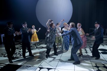 Joukko ihmisiä, joista osa on pukeutunut värikkäisiin mekkoihin ja osa pukuihin ja hattuihin, tanssii tarmokkaasti kuviollisella lavalla, jonka taustalla on suuri valkoinen ympyrä dramaattisessa valaistuksessa.