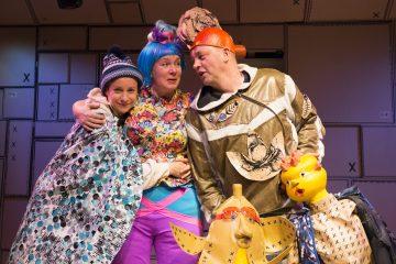 Kolme näyttelijää värikkäissä, omituisissa puvuissa seisoo yhdessä lavalla, hymyilee ja poseeraa. Lavasteet heidän takanaan on koristeltu pahvilaatikoilla. Etualalla näkyy kaksi keltaista ankka-rekvisiittaa.