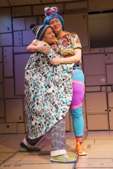 Kaksi värikkäisiin, omituisiin pukuihin pukeutunutta ihmistä syleilee lämpimästi lavalla. Toisella on kuvioitu kaapu ja pipo, kun taas toisella on kirkkaan siniset hiukset, kukkapaita ja värikkäät legginsit. Taustalla on pahvilaatikoita.