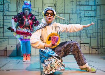 Lavalla esiintyy kaksi näyttelijää värikkäissä, omituisissa puvuissa. Edessä oleva henkilö poseeraa energisesti, hänellä on silmälasit päässään, hänellä on kädessään lelusekoitin ja kuviollinen viitta, kun taas toinen seisoo takana kirkkaisiin, leikkisiin vaatteisiin ja siniseen peruukkiin pukeutuneena.