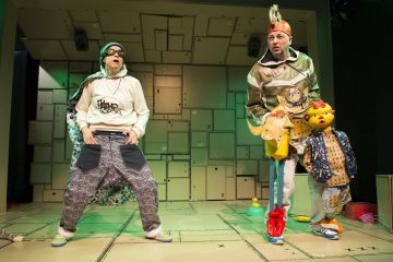 Kaksi esiintyjää seisoo lavalla, jonka taustalla on pinottuja pahvilaatikoita. Toisella on päällään huppari, aurinkolasit ja pussihousut, toisella on hienostunut puku, jossa on kypärä ja värikkäitä rekvisiittaa. Kohtaus vaikuttaa teatraaliselta ja leikkisältä.