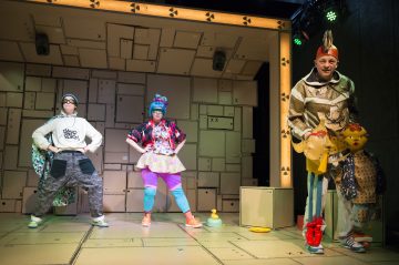 Kolme esiintyjää värikkäissä, omituisissa puvuissa poseeraavat leikkisästi lavalla, jonka taustalla on pahvilaatikoita ja kirkas valaistus, mikä luo omituisen teatteritunnelman.