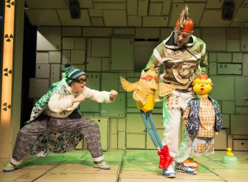 Kaksi värikkäisiin, omituisiin pukuihin pukeutunutta näyttelijää esiintyy energisesti lavalla; toinen imitoi lyöntiä, kun taas toisella on kädessään kaksi suurta, omituista nukkea. Lavastusta koristavat geometriset, teollisen tyylin paneelit.