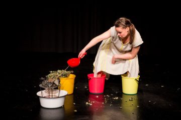 Valkoiseen mekkoon pukeutunut nainen kumartuu kolmen värikkään ämpärin yli tummalla näyttämöllä ja siirtää punaisella kauhalla lehtijätteitä siivilästä ämpäriin. Joitakin lehtiä on hajallaan lattialla.