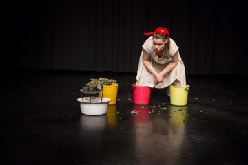 Valkoiseen mekkoon ja punaiseen hattuun pukeutunut nainen kyyristelee kolmen värikkään ämpärin välissä tummalla näyttämöllä, jossa lehdet ovat hajallaan maassa ja kori lepää valkoisessa ämpärissä.