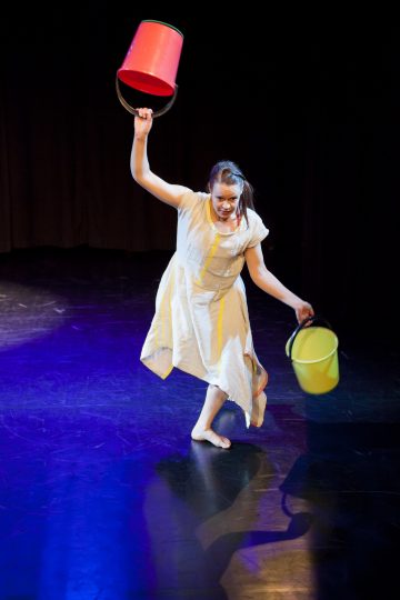 Valkoiseen mekkoon keltaisilla raidoilla pukeutunut paljasjalkainen nainen tanssii lavalla pitäen päänsä yläpuolella punaista ämpäriä ja toisessa kädessään keltaista ämpäriä värikkäässä valaistuksessa.