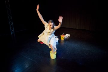 Valkoiseen mekkoon pukeutunut nainen astuu yhdellä jalalla keltaiseen ämpäriin ja nostaa molemmat kädet ylös tummalla näyttämöllä. Taustalla näkyy kaksi muuta ämpäriä ja pyyhe.