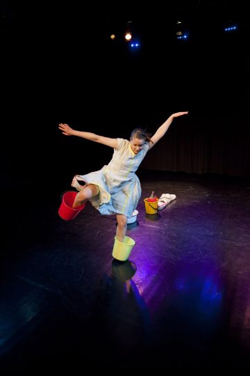 Valkoiseen mekkoon pukeutunut tyttö tasapainoilee lavalla yhdellä jalalla, kädet ojennettuina ja toinen jalka keltaisen ämpärin sisällä. Toinen punainen ämpäri on lähellä, ja näyttämön valot heijastavat värikkäitä heijastuksia lattiaan.