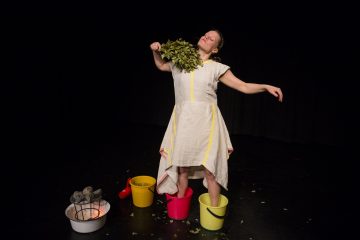 Valkoiseen mekkoon pukeutunut nainen seisoo jalat kahdessa muoviämpärissä ja pitää lehtien oksia kaulassaan. Lähistöllä on musta tausta, jossa on metallinen ämpäri, jossa on kiviä, ja toinen ämpäri, jossa on vettä.