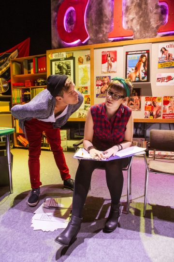 Pipoon pukeutunut mies nojaa kohti istuvaa silmälasipäistä naista, joka kirjoittaa papereihin. He ovat värikkäässä huoneessa, jonka seinällä heidän takanaan on lehtiä ja julisteita.