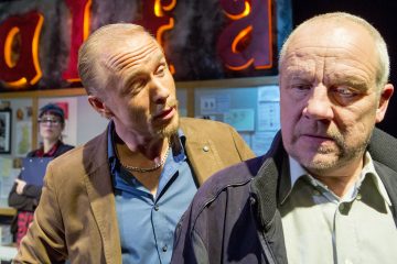 Kaksi miestä seisoo lähekkäin, toinen puhuu tiiviisti toiselle, joka katsoo vakavana poispäin. Taustalla seisoo nainen, jolla on silmälasit ja leikepöytä. Heidän takanaan olevalla seinällä näkyy osittain kirkkaan oransseja kirjaimia.