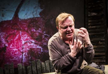 Vaaleatukkainen henkilö, jolla on tuskallinen ilme, puristaa käsiään ja näyttää huutavan tai itkevän lavalla. Taustalla on voimakasta punaista ja tummaa abstraktia kuvamateriaalia, mikä viittaa dramaattiseen kohtaukseen.