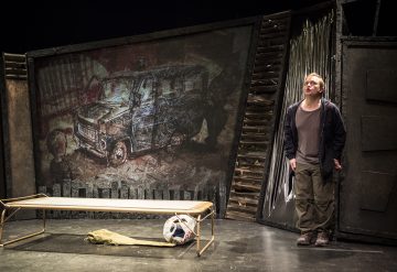 Henkilö seisoo hämärästi valaistulla näyttämöllä yksinkertaisen penkin vieressä, ja lattialla on kulunut jalkapallo ja kangas. Heidän takanaan on seinämaalaus, joka kuvaa vaurioitunutta pakettiautoa ja kaoottista kohtausta. Lavastuksessa on teollisia ja ahdistuneita elementtejä.