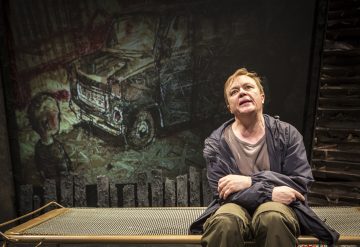 Henkilö istuu pinnasängyllä kädet ristissä ja katsoo ylöspäin ahdistunut ilme kasvoillaan. Heidän takanaan on tumma, taiteellinen tausta, jossa on piirros kuorma-autosta ja lapsen näköinen hahmo. Asetelma vaikuttaa dramaattiselta ja jännittyneeltä.