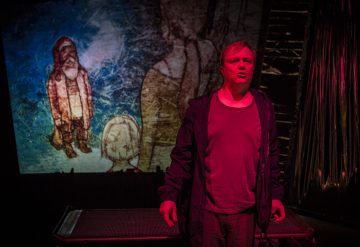 Henkilö seisoo punaisen näyttämövalaistuksen alla puhumassa tai esiintymässä, ja hänen takanaan olevalle kuvaruudulle heijastetaan kahden hahmon kuvitettu kohtaus. Asetelma viittaa teatteri- tai draamaesitykseen.