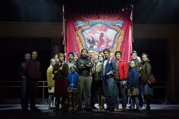 Joukko työväen vaatteisiin pukeutuneita ihmisiä seisoo yhdessä lavalla suuren punaisen Durhamin kaivostyöläisten yhdistyksen banderollin edessä, joka on dramaattisesti valaistu ja herättää solidaarisuutta ja päättäväisyyttä.