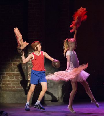 Nyrkkeilyvarusteisiin pukeutunut poika ja tutuihin pukeutunut tyttö tanssivat yhdessä lavalla, kummallakin sulkatuke kädessään, ja taustalla on dramaattinen valaistus ja tiiliseinä.