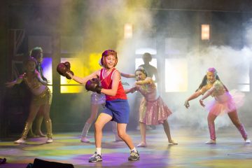 Nyrkkeilyhanskoihin, shortseihin ja toppiin pukeutunut lapsi tanssii lavalla hymyillen värikkäisiin, kimalteleviin pukuihin ja tutuihin pukeutuneiden tyttöjen ympäröimänä, kirkkaiden lavalamppujen ja savun loisteessa taustalla.