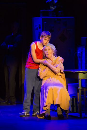 Nuori poika, jolla on punainen toppi, lohduttaa keltaiseen mekkoon ja huiviin pukeutunutta iäkästä naista hämärästi valaistulla näyttämöllä, mikä viittaa teatteriesityksen dramaattiseen kohtaukseen.