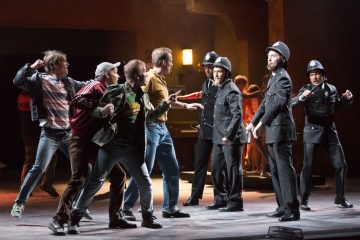 Ryhmä miehiä kohtaa teatterin lavalla univormupukuisia poliiseja, ja heidän ruumiinkielensä on jännittynyt ja heidän asenteensa dramaattisia, mikä viittaa kiihkeään tai tunteikkaaseen esityskohtaukseen.