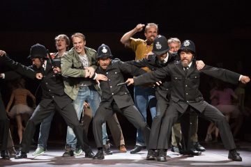 Ryhmä näyttelijöitä näyttämöllä, joista jotkut ovat pukeutuneet brittiläisiksi poliiseiksi ja poseeraavat dramaattisesti, kun taas toiset satunnaisiin vaatteisiin pukeutuneet näyttelijät elehtivät tarmokkaasti heidän takanaan näyttämövalaistuksen alla.