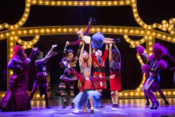 Vilkas sirkusteemainen lavaesitys, jossa esiintyjät ovat pukeutuneet värikkäisiin asuihin, kuten tutuihin ja silinterihattuihin, ja taustalla on kirkkaat telttavalot, jotka hahmottavat suurta rakennusta.