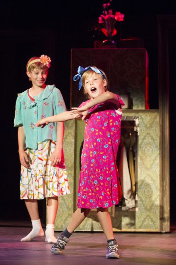 Lavalla esiintyy kaksi lasta värikkäissä mekoissa; toinen sinisessä yläosassa ja kuviollisessa hameessa hymyilee, kun taas toinen kirkkaan vaaleanpunaisessa mekossa ja sinisessä otsanauhassa tanssii tarmokkaasti parrasvaloissa. Taustalla on kukka-asetelma.