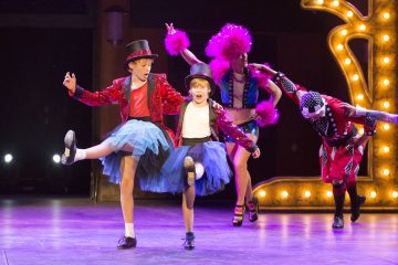 Kaksi sinisiin tutuihin, punaisiin paljettitakkeihin ja silinterihattuihin pukeutunutta lasta tanssii lavalla vilkasta tanssia, ja heidän seuranaan on kaksi aikuista värikkäissä teatteripuvuissa ja peruukissa kirkkaiden valojen alla.