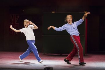 Nuori poika ja nainen, molemmat urheiluvaatteissa, tanssivat lavalla vilkasta tanssia ilmeikkäissä asennoissa. Taustalla on haalea Britannian lippu ja näyttämövalaistus.