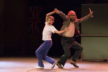 Valkoiseen paitaan ja sinisiin housuihin pukeutunut nuori poika tanssii tarmokkaasti silmälasipäisen ja takkiin pukeutuneen vanhemman miehen kanssa. Molemmilla on ilmeikkäät asennot, ja taustalla näkyy Britannian lippu.