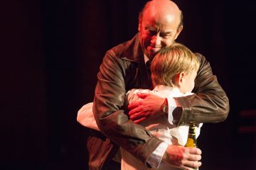 Nahkatakkinen mies halajaa lämpimästi nuorta poikaa, jolla on pullo kädessään. Molempia valaisee pehmeä, lämmin valaistus tummaa taustaa vasten.