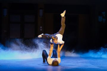 Kaksi valkoisiin paitoihin ja tummiin housuihin pukeutunutta esiintyjää tekee lavalla akrobatiaa; toinen makaa lattialla ja nostaa toista ylösalaisin ilmaan, ja taustalla on lavasavua ja sininen valaistus.