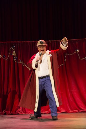 Punaiseen kaapuun, kruunuun ja sinisiin housuihin pukeutunut vanhempi mies pitelee mikrofonia ja elehtii tarmokkaasti lavalla punaisilla verhoilla, joita koristavat jousivalot.