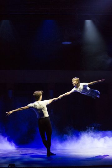 Kaksi esiintyjää lavalla, toinen pitää toista kädestä kiinni. Toinen esiintyjä roikkuu vaakatasossa ilmassa, ikään kuin lentäen. Siniset näyttämövalot ja savu luovat dramaattisen tunnelman.