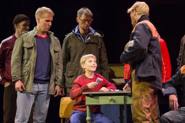 Nuori poika istuu pienen pöydän ääressä hymyillen ja kortteja kädessään, ja hänen ympärillään on neljä seisovaa ja yksi istuva mies, jotka kaikki käyvät vilkasta vuorovaikutusta teatterin lavalla.