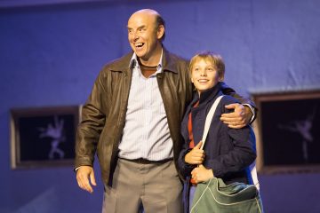 Hymyilevä mies, jonka käsi on pojan ympärillä, joka myös hymyilee ja jolla on säkki kädessään, seisovat yhdessä lavalla, jonka taustalla on kehystettyjä kuvia seinällä.