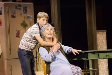 Nuori raitapaitainen poika halaa keittiön pöydän ääressä istuvaa iäkästä naista. Nainen, jolla on sininen kaapu ja päähuivi, katsoo ylöspäin ja hymyilee. Taustalla näkyy jääkaappi, jossa on magneetteja.