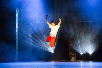 Valkoiseen paitaan ja punaisiin housuihin pukeutunut nuori hyppii energisesti lavalla kädet ylhäällä, ja taustalla on dramaattinen sininen valaistus ja valonheittimet, jotka luovat säteitä.