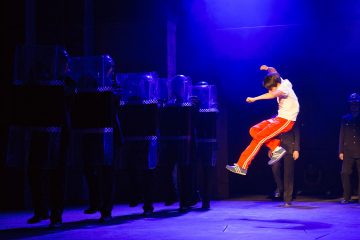 Punaisiin housuihin ja valkoiseen paitaan pukeutunut nuori hyppää ilmassa mellakkasuojia pitelevien poliisien edessä pimeässä, sinisellä ja violetilla valaistussa lavastuksessa.