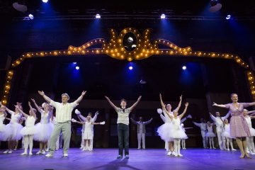 Ryhmä esiintyjiä, joihin kuuluu valkoisiin balettiasuihin pukeutuneita lapsia ja pukuihin pukeutuneita aikuisia, seisoo lavalla kädet ojennettuina valaistun kaaren alla teatteriesityksen aikana.