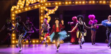 Värikkäisiin, sirkusaiheisiin pukuihin pukeutuneet esiintyjät tanssivat energisesti lavalla, ja taustalla on suuri, valaistu teltta, joka luo eloisan ja juhlavan tunnelman.