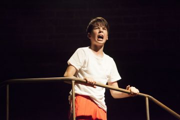 Valkoiseen t-paitaan ja oransseihin housuihin pukeutunut nuori henkilö seisoo metallikaiteella, tarttuu siihen tiukasti ja huutaa, ja hänen ilmeensä on intensiivinen dramaattisessa näyttämövalaistuksessa.