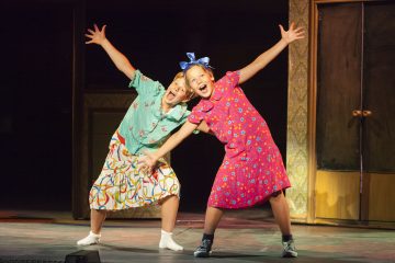 Kaksi värikkäisiin mekkoihin pukeutunutta nuorta tyttöä seisoo lavalla leveästi hymyillen, kädet ojennettuina ja jalat levällään, ja he vaikuttavat iloisilta ja energisiltä teatteriesityksen aikana.