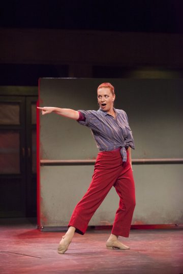 Lavalla esiintyy raidalliseen paitaan ja punaisiin housuihin pukeutunut nainen, joka osoittaa dramaattisesti sivulle toinen käsi ojennettuna, keskellä tanssia, ilmeikkäät kasvot ja teatraalinen valaistus.