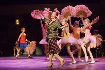 Nainen tanssii keskellä näyttämöä, ja hänen takanaan on nuoria tyttöjä tutuissa, joilla on sulkatäytteisiä viuhkoja. Vasemmalla puolella seisoo nyrkkeilyhanskat kädessään poika, jolla on toppi ja shortsit. Tausta on hämärästi valaistu, mikä viittaa teatteriesitykseen.