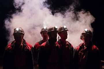 Viisi kaivosmiestä, joilla on kypärä ja otsalamppu, seisovat yhdessä dramaattisen valaistuksen valaisemina, ja heidän takanaan pimeässä maisemassa on sakeaa savua tai sumua.